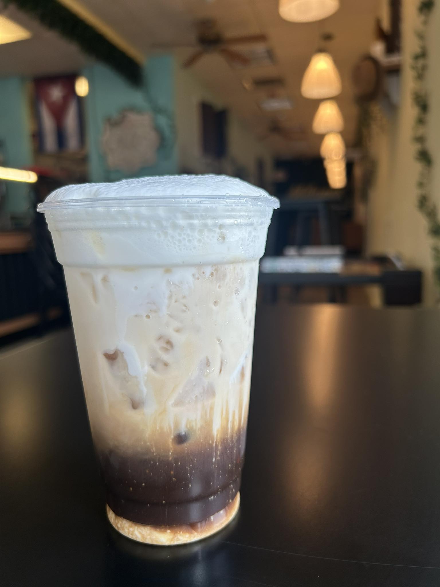 Tall layered iced cafe con leche in a clear cup on the Milanes Cuban Corner counter, dining room and Cuban flag visible behind