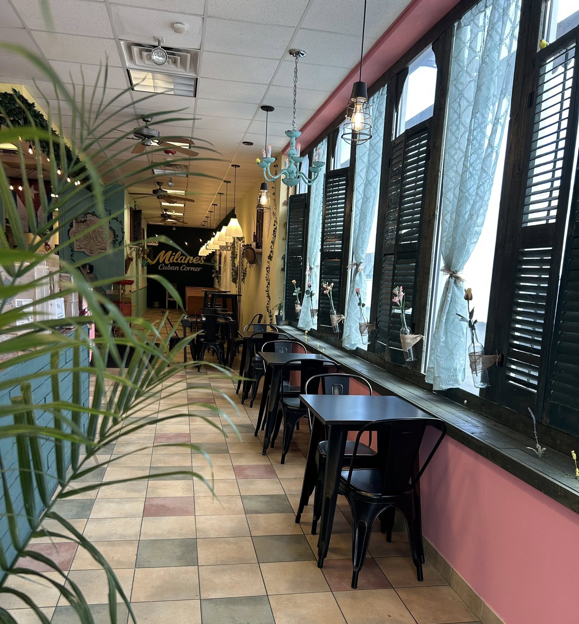 Sunlit interior of Milanes Cuban Corner: row of black bistro tables along blue plantation-shutter windows under brass chandeliers